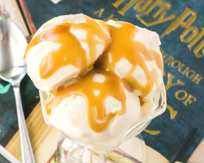overhead view of three scoops of Ninja Creami Butterbeer ice cream served in a glass dessert cup, topped with butterscotch sauce. A Harry Potter book title is in the background, and a spoon rests off to the side.
