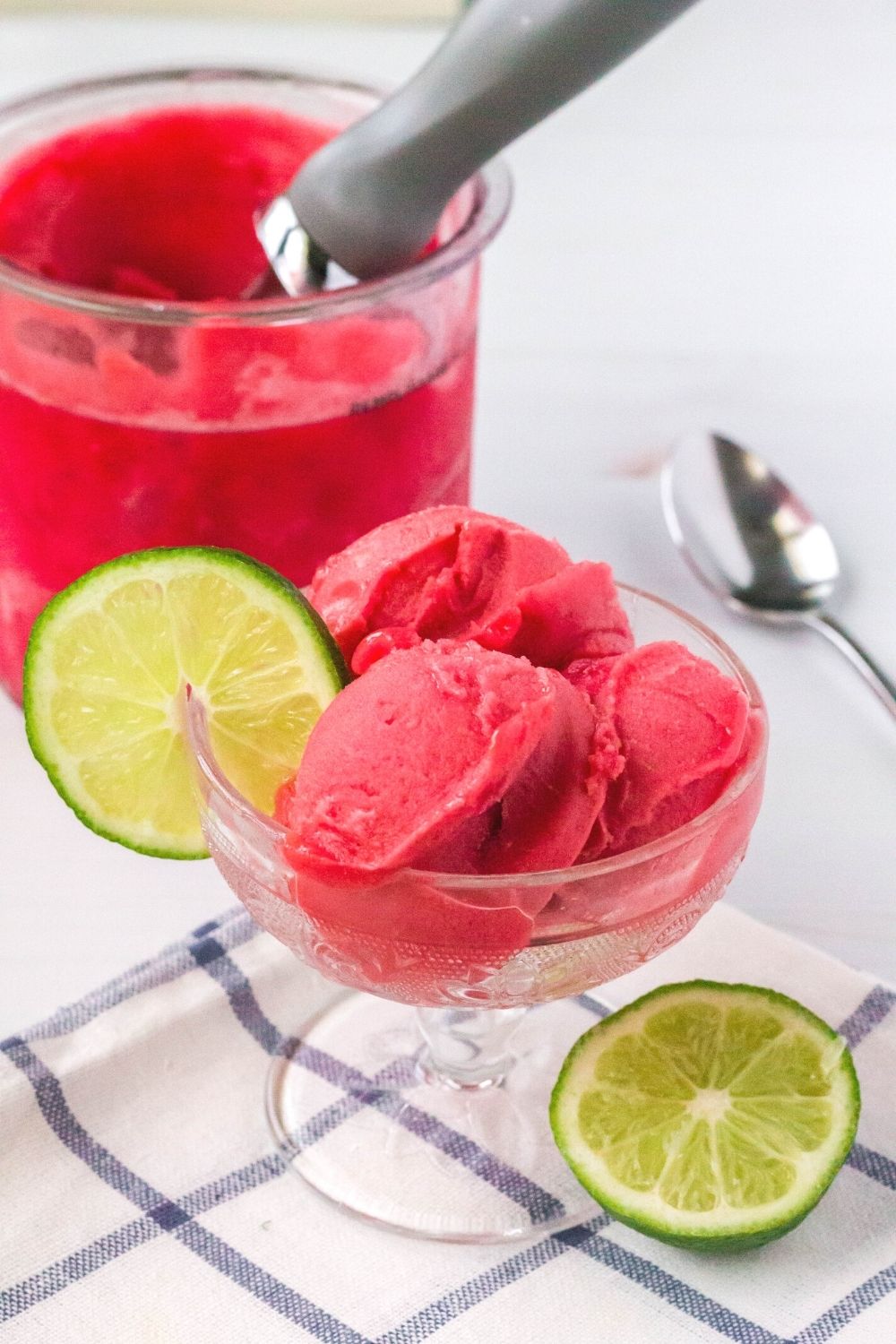 A glass dessert cup serving scoops of Ninja Creami cherry limeade sorbet is in the foreground; and a Ninja Creami pint of the remaining sorbet is in the background.