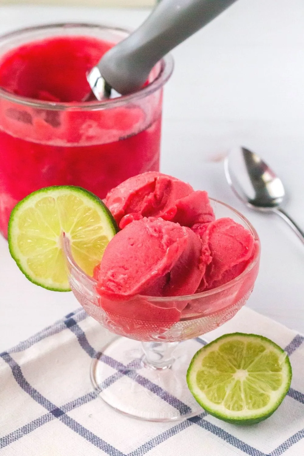 A glass dessert cup serving scoops of Ninja Creami cherry limeade sorbet is in the foreground; and a Ninja Creami pint of the remaining sorbet is in the background.