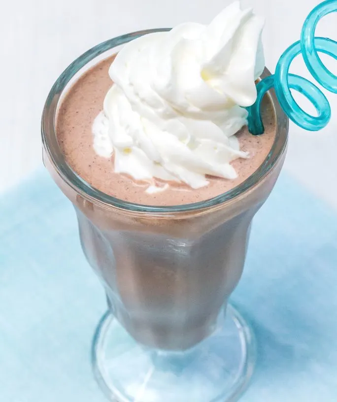 Ninja Creami chocolate milkshake in an ice cream glass. The milkshake is topped with a swirl of whipped cream and a blue spiral straw.