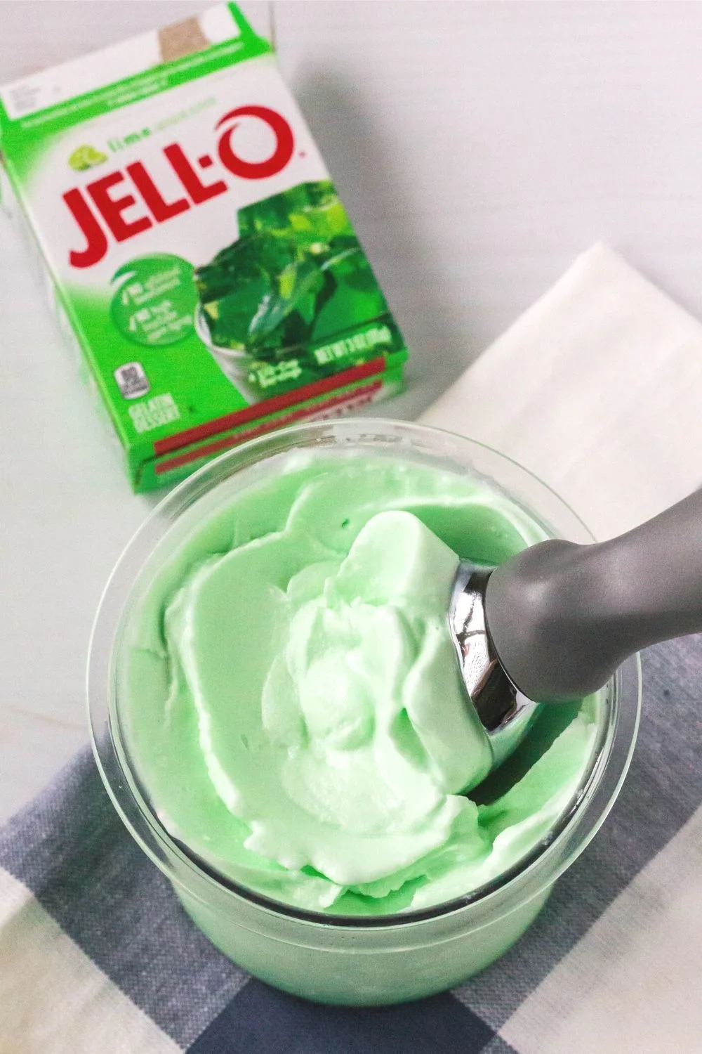 overhead view of a Ninja Creami pint container of lime sherbet, with an ice cream scoop in it. An empty box of lime Jell-O gelatin mix is next to the pint.