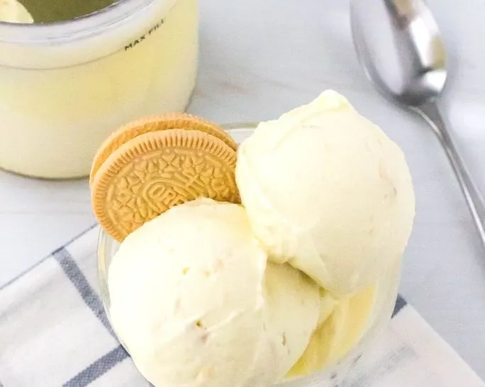 a glass serving dish containing scoops of Ninja Creami lemon ice cream, with the remaining pint and a spoon in the background.
