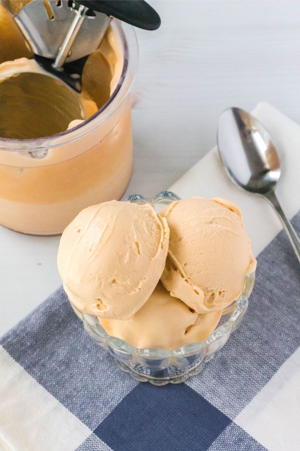 a glass dessert cup serving three scoops of Ninja Creami butterscotch pudding ice cream is in the foreground. A pint container and spoon are in the background.