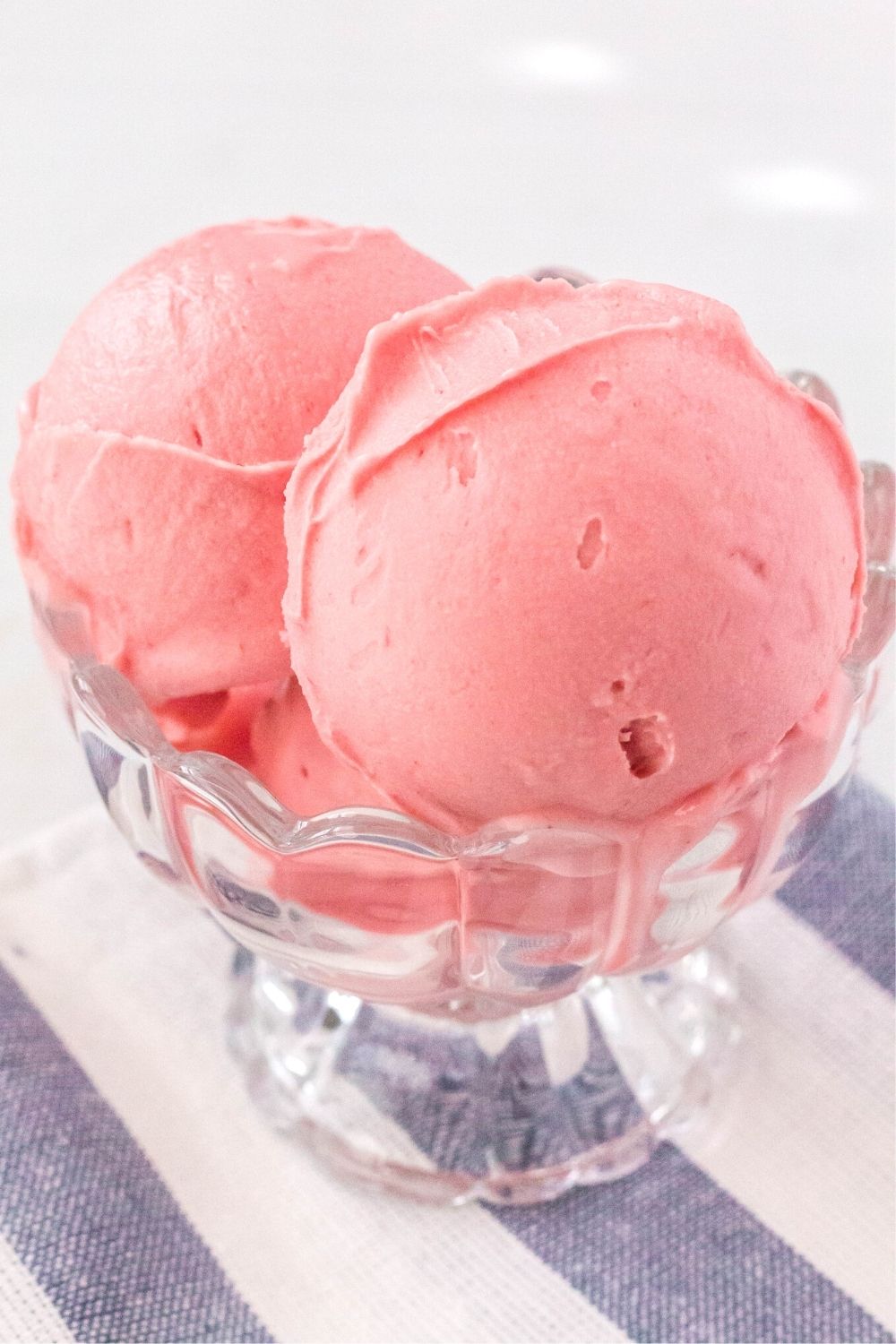 side view of scoops of cherry cheesecake ice cream served in a glass ice cream dish, atop a blue and white striped napkin.