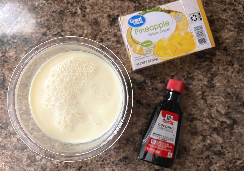 a box of pineapple gelatin dessert and a bottle of coconut extract are next to a Ninja Creami pint with pina colada ice cream base in it.