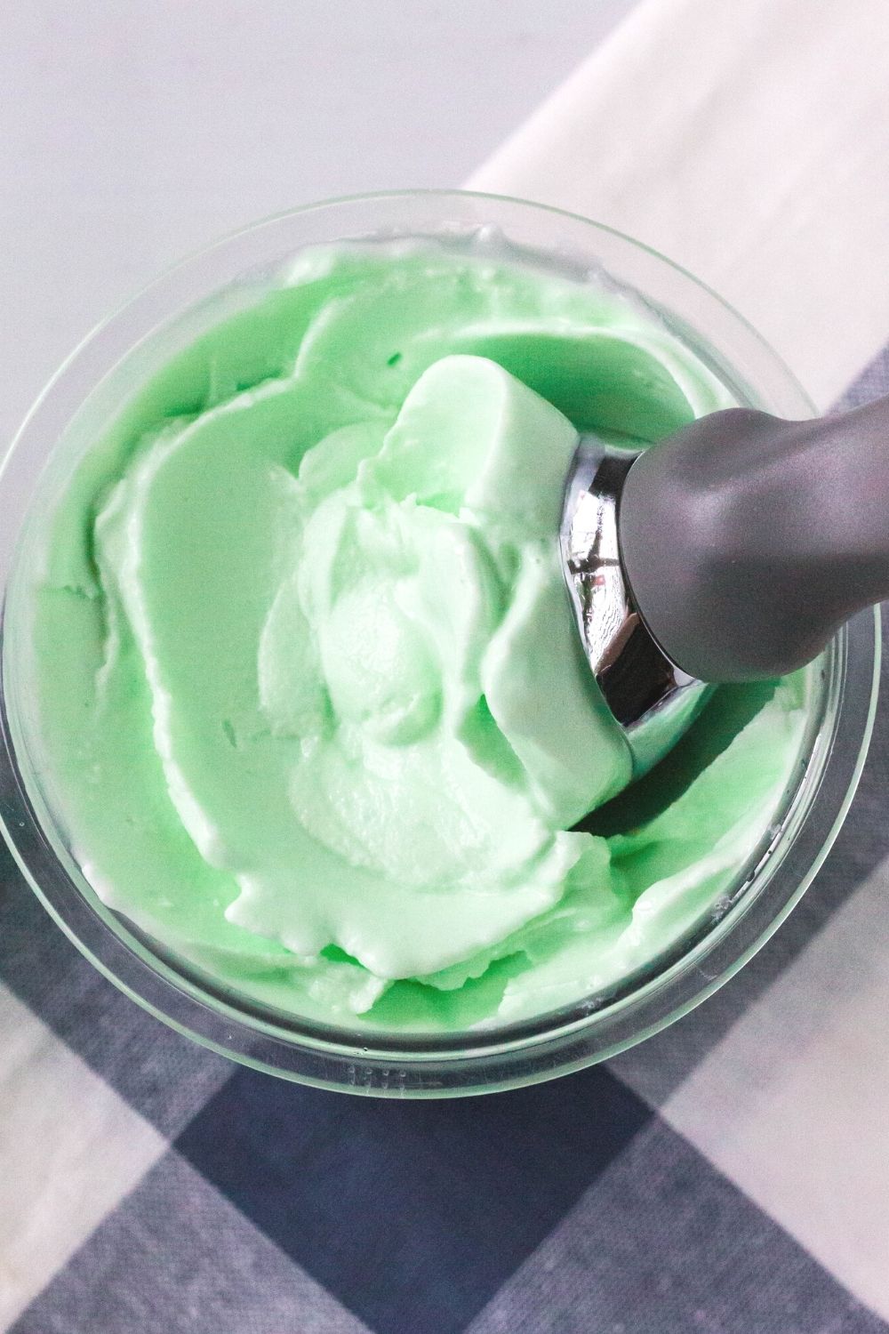 overhead view of green lime sherbet in a ninja creami pint container, atop a blue and white checked napkin. An ice cream scoop is in the sherbet.