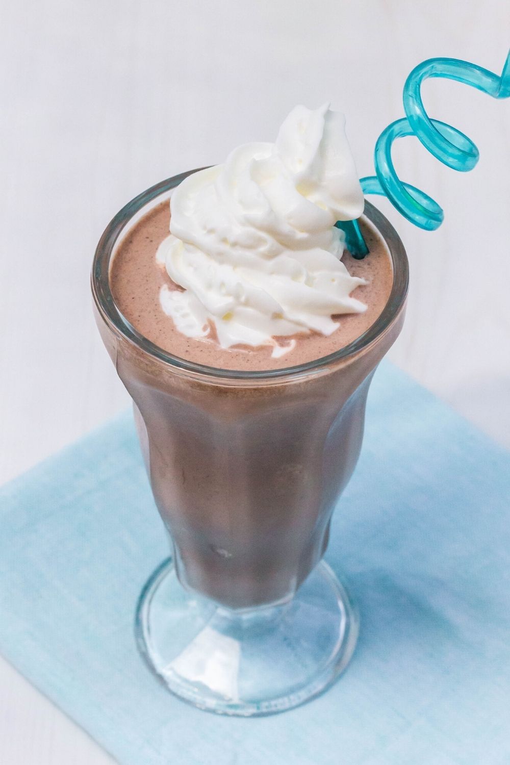 an ice cream milkshake glass is filled with a Ninja Creami milkshake and topped with whipped cream. A blue plastic swirly straw is in the glass.