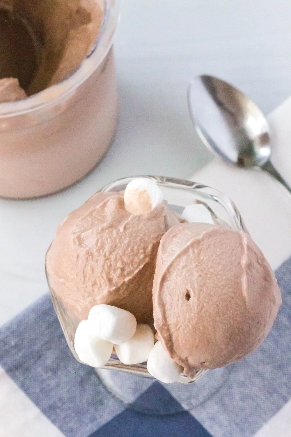overhead view of scoops of frozen hot cocoa ice cream in a dessert cup, topped with mini marshmallows. A Ninja Creami pint container is in the background, along with a spoon.