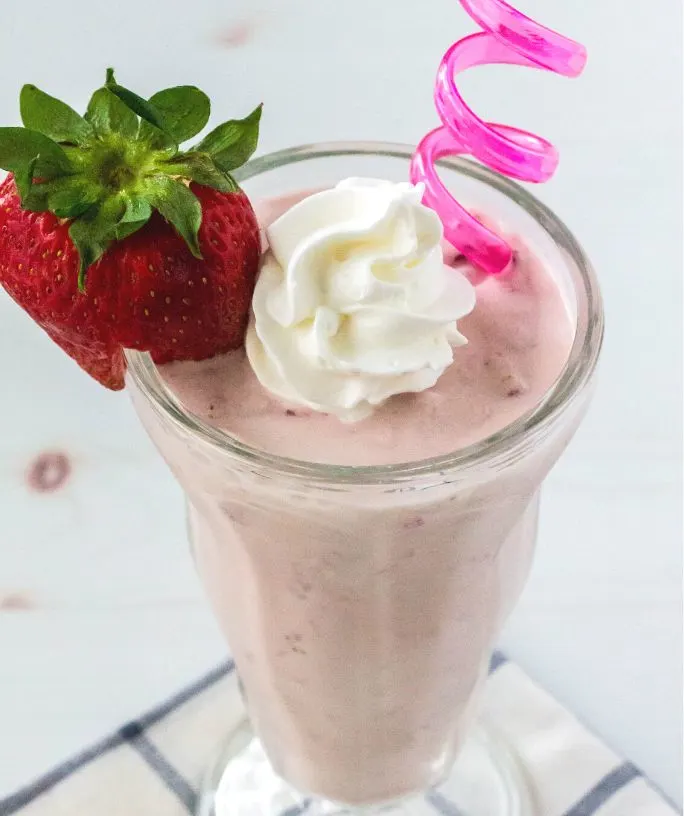 a glass of Ninja Creami strawberry milkshake is garnished with a fresh strawberry, dollop of whipped cream, and served with a pink straw.