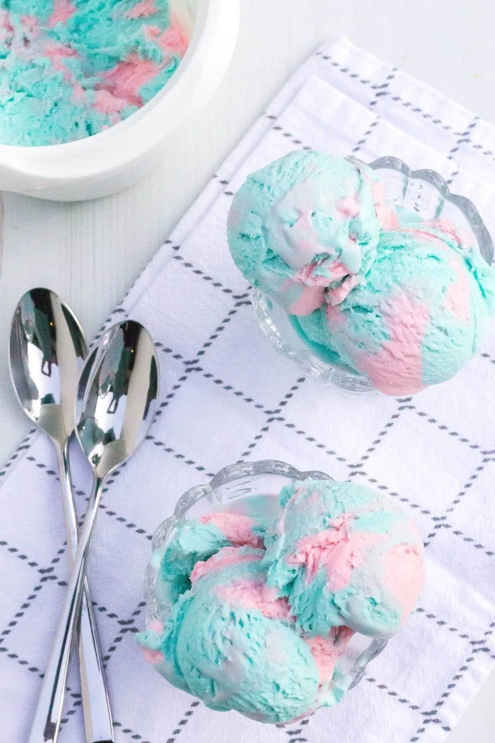 overhead view of two dessert cups filled with scoops of cotton candy ice cream made without an ice cream machine. The ice cream container and two spoons are in the background.