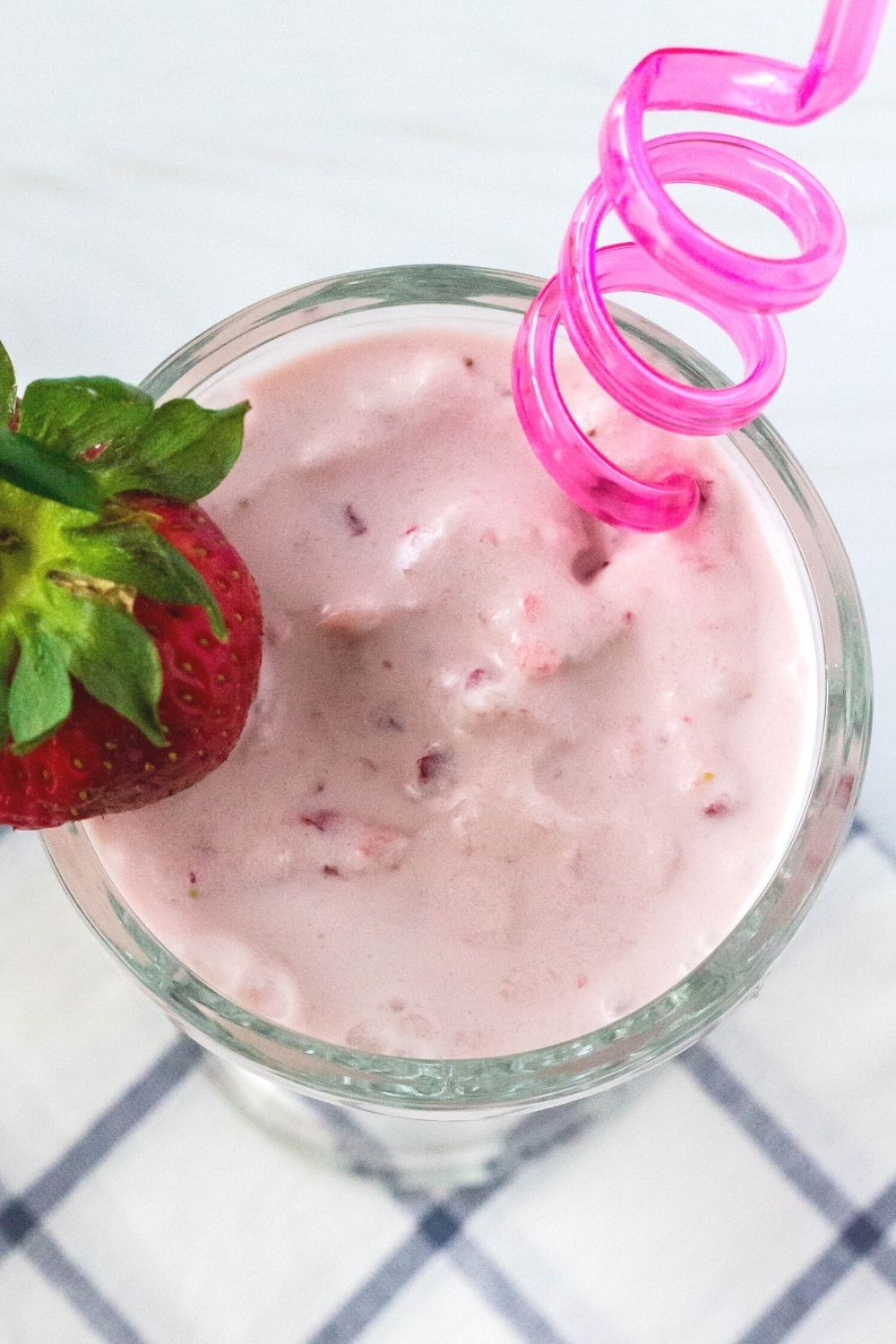 overhead view of a glass filled with strawberry milkshake made in the Ninja Creami machine, garnished with a fresh strawberry and served with a pink swirly straw.