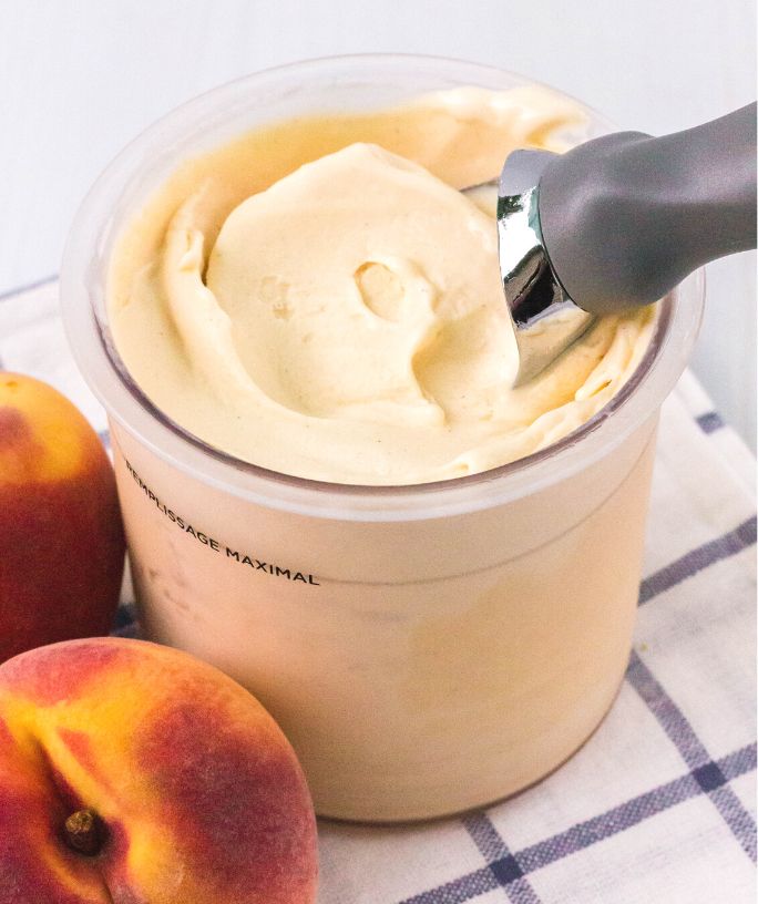 Ninja Creami peach ice cream in a pint with an ice cream scoop. Two fresh peaches are next to the container.