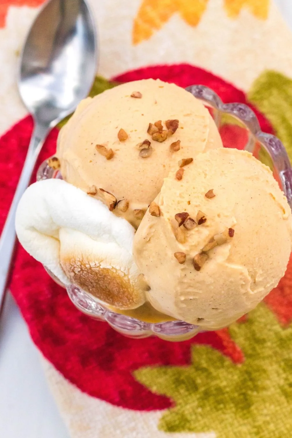 overhead view of a dessert cup filled with sweet potato ice cream, sprinkled with pecans and garnished with a marshmallow. A spoon is next to the dish.