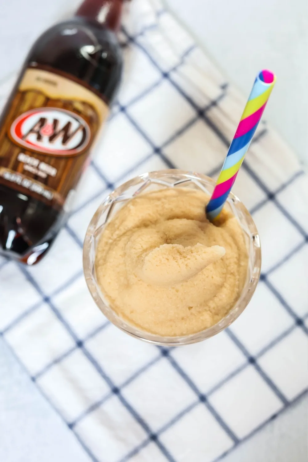 overhead view of the surface of a slushy made with root beer. A straw is in the slushy and a bottle of root beer is next to it.