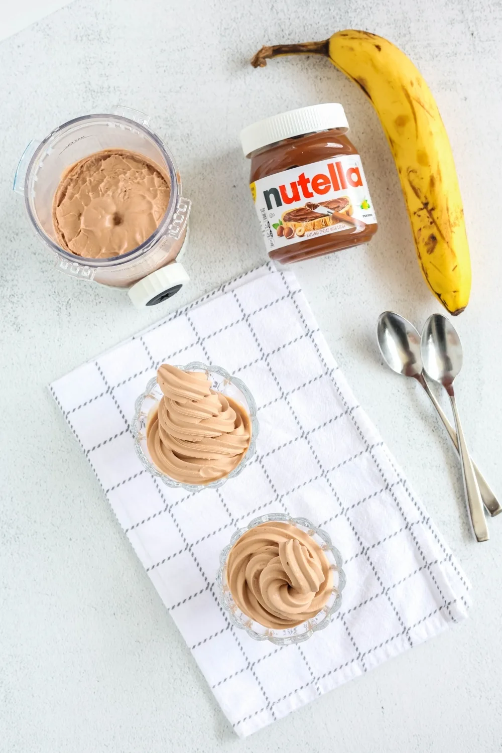 overhead view of two glass dishes of Nutella and banana frozen yogurt. Two spoons, a banana, a jar of chocolate hazelnut spread, and a Ninja Creami pint container are scattered around the dishes.