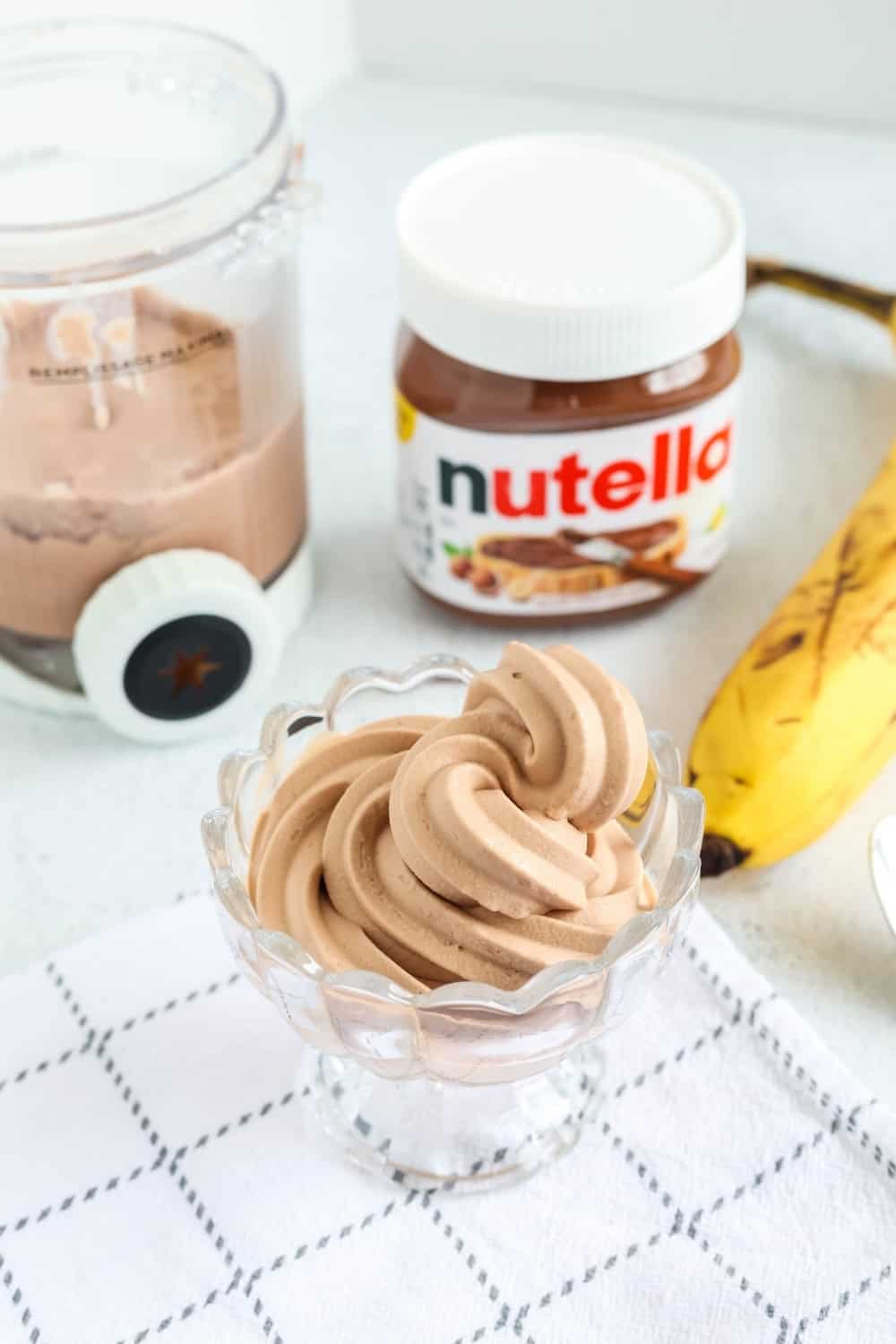 Ninja Creami Swirl banana Nutella frozen yogurt served in a glass dish. The remaining pint of FroYo is in the background, along with a jar of Nutella and a ripe banana.