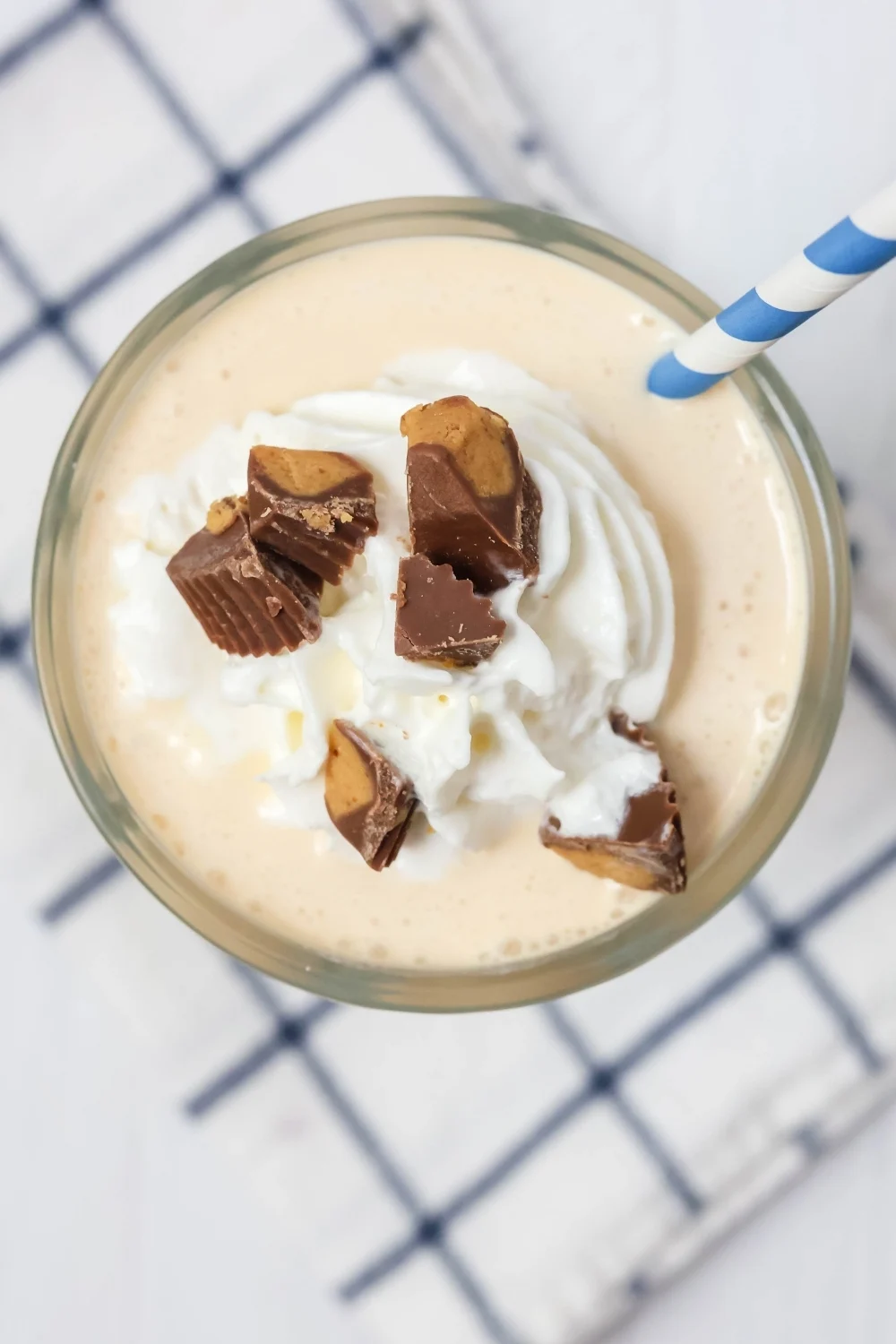 overhead view of ninja creami peanut butter milkshake in a glass, topped with reese's peanut butter cup pieces and whipped cream, with a blue and white straw.
