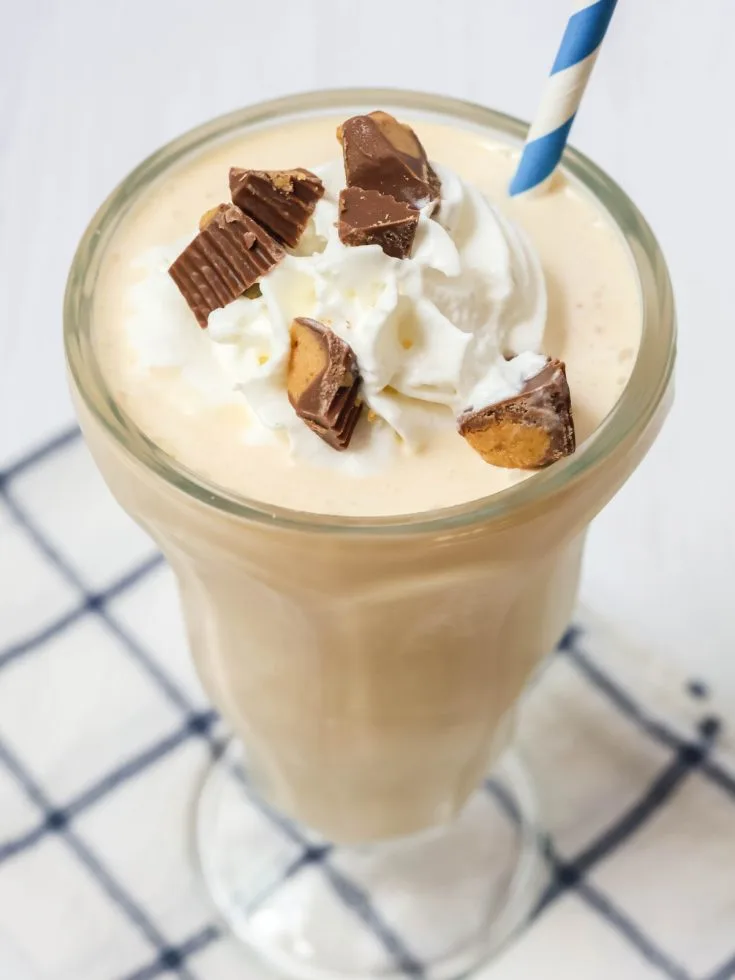 Ninja Creami peanut butter milkshake served in a malt glass, topped with whipped cream and chopped peanut butter cups. A blue and white straw is in the glass.