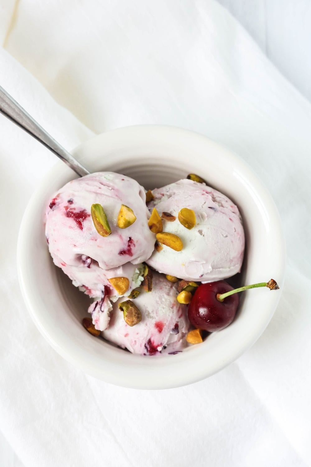 overhead view of a white bowl with three scoops of cherry pistachio ice cream made in the Ninja Creami machine. The ice cream is garnished with fresh pistachios and a fresh cherry.