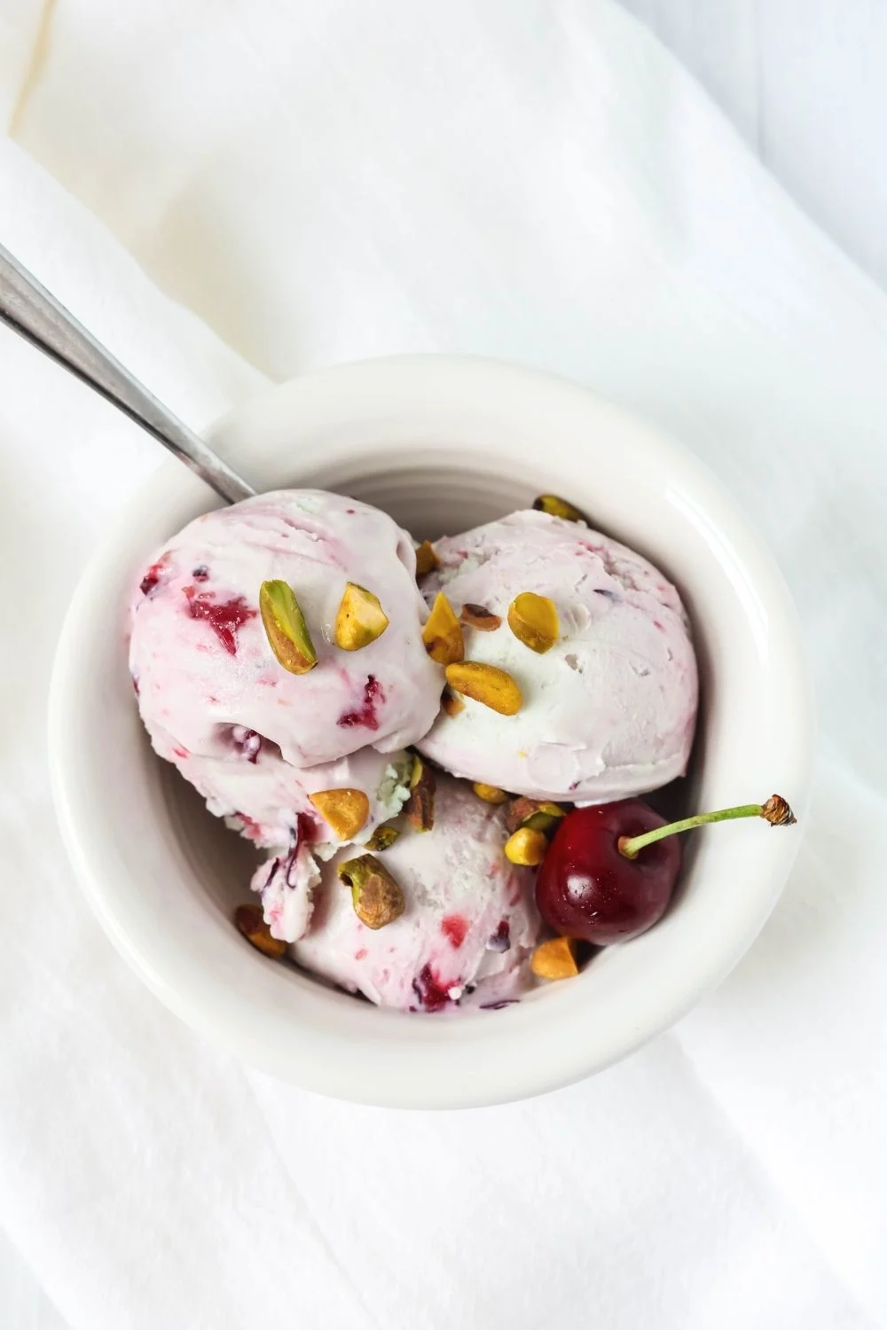 overhead view of a white bowl with three scoops of cherry pistachio ice cream made in the Ninja Creami machine. The ice cream is garnished with fresh pistachios and a fresh cherry.