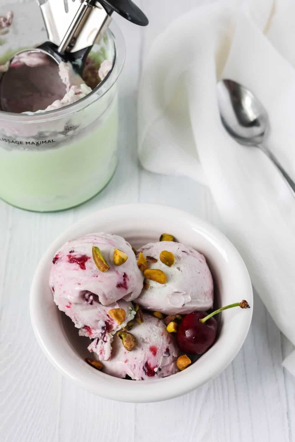 a white bowl serves scoops of pistachio and cherry ice cream. A ninja creami pint of remaining ice cream is in the background, along with a spoon.