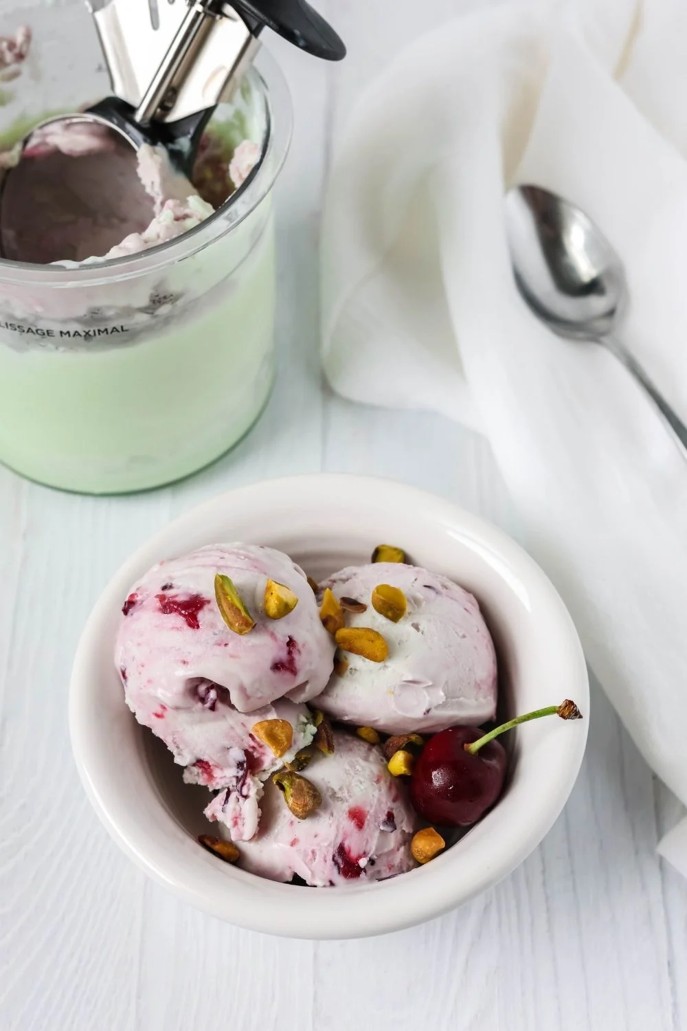 a white bowl serves scoops of pistachio and cherry ice cream. A ninja creami pint of remaining ice cream is in the background, along with a spoon.