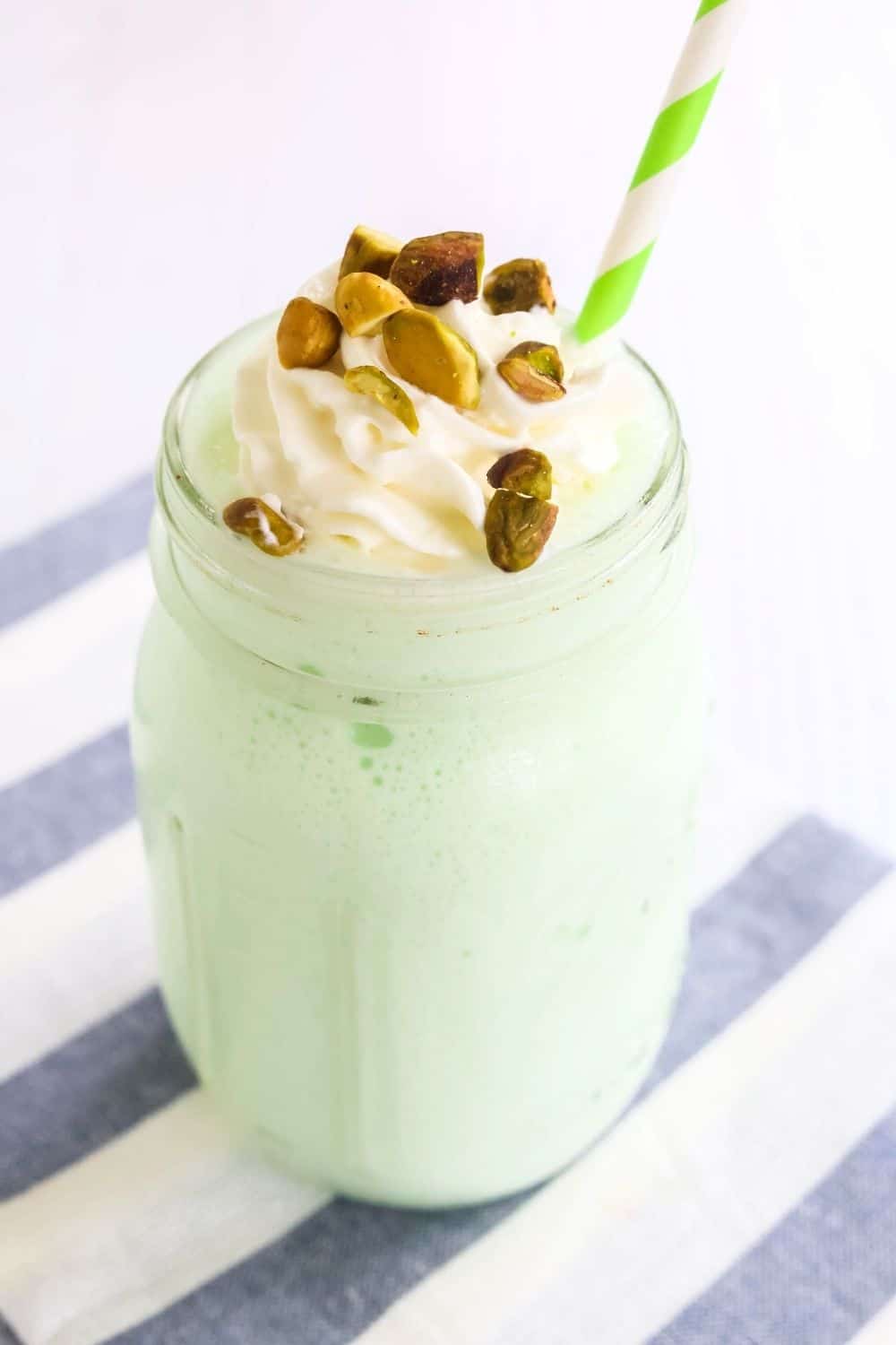 glass of pistachio milkshake made in the Ninja Slushi machine. A green and white straw is in the shake, and it's topped with nuts and whipped cream.