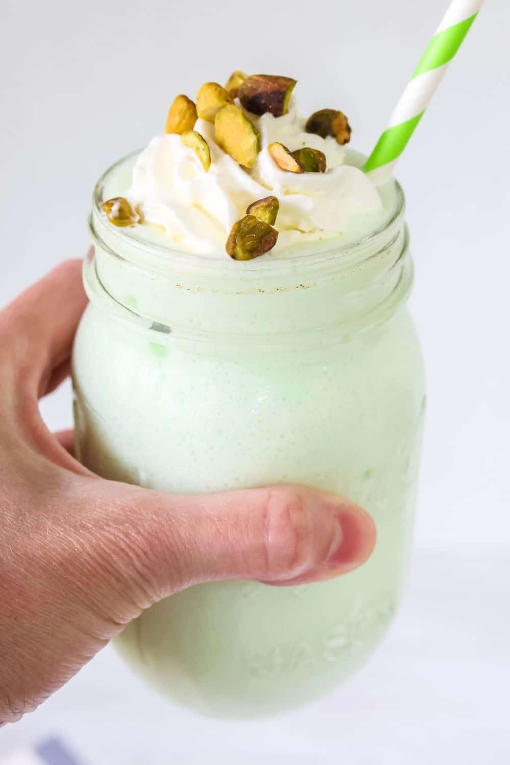 a woman's hand holds a mason jar filled with pistachio pudding milkshake made in the Ninja Slushi machine, topped with whipped cream and pistachios.