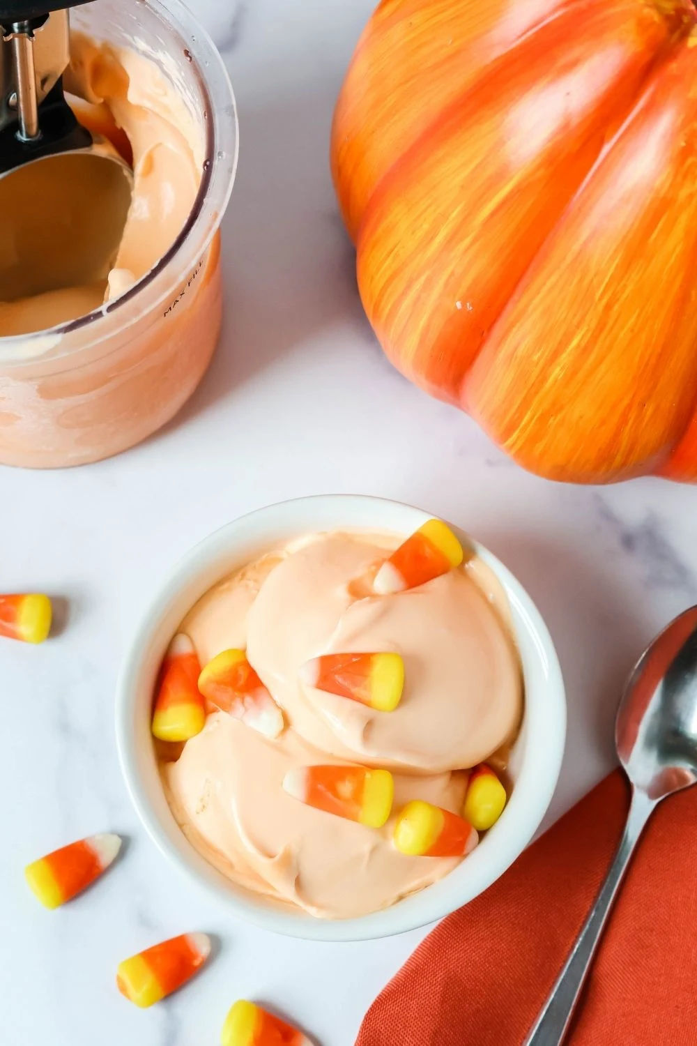overhead view of a white bowl with scoops of candy corn ice cream in it. A Ninja Creami pint of remaining ice cream is in the background, along with a decorative pumpkin and candy corn candies.