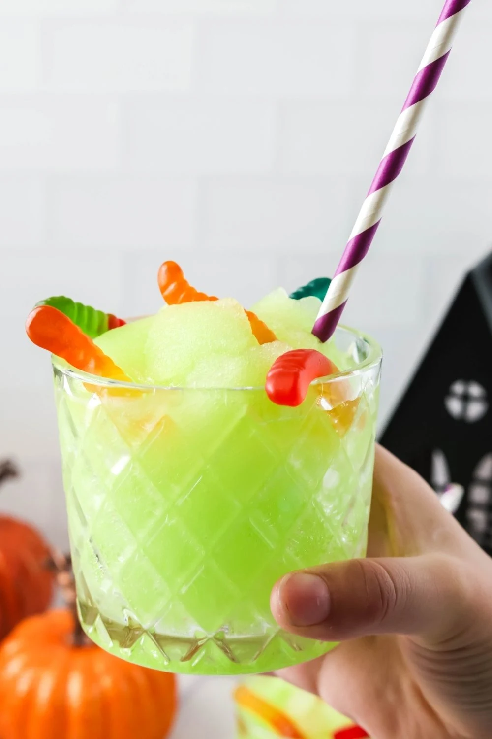 a hand holds a glass of Ninja Slushi bright green Halloween slush, garnished with gummy worms and a purple and white striped straw.