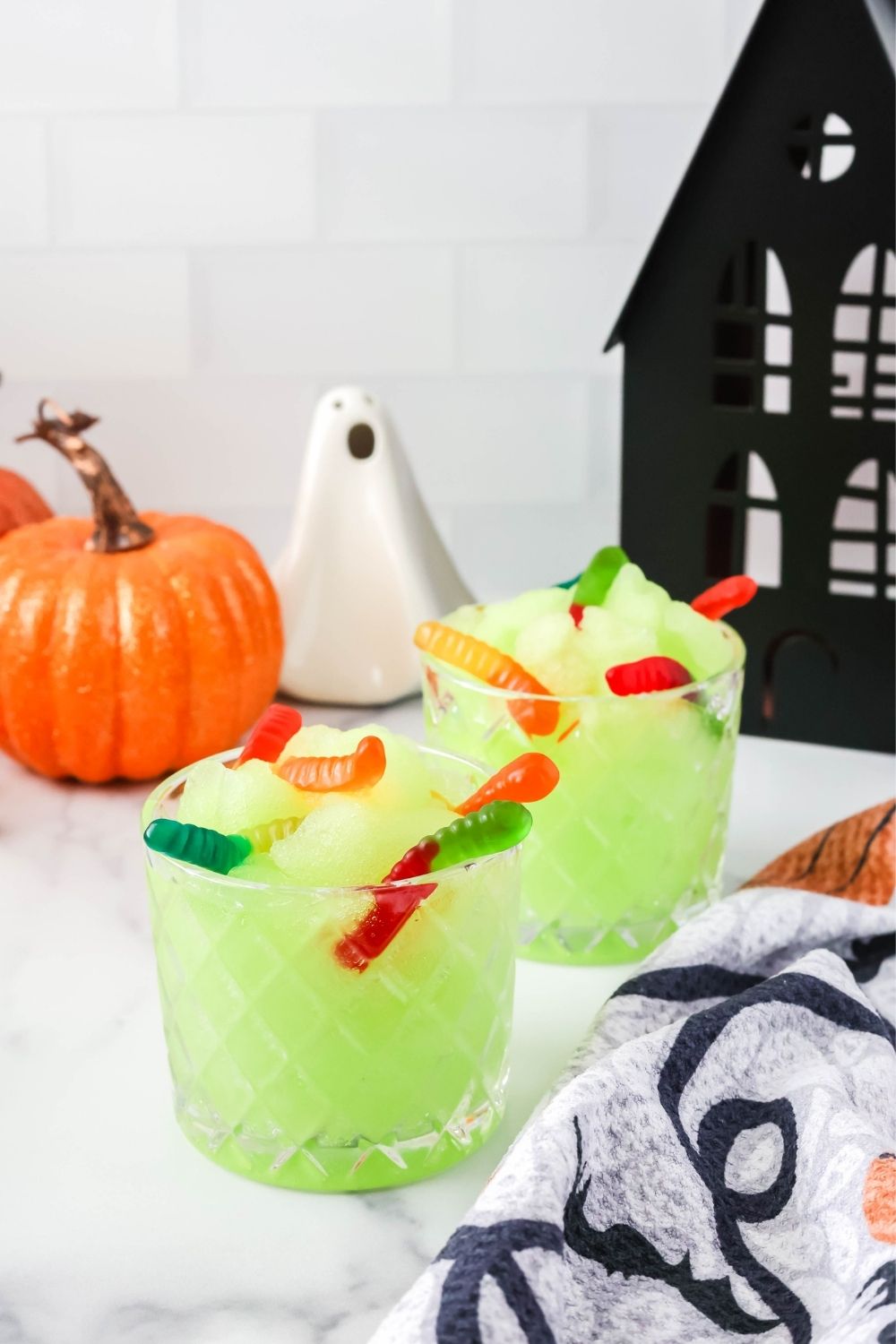 two glasses of Ninja Slushi machine Halloween slush, garnished with gummy worms. Halloween decor is around the glasses of green slush.