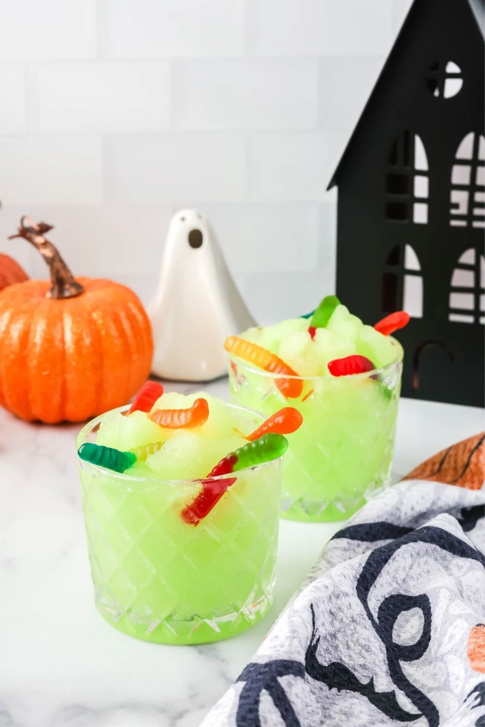 two glasses of Ninja Slushi machine Halloween slush, garnished with gummy worms. Halloween decor is around the glasses of green slush.