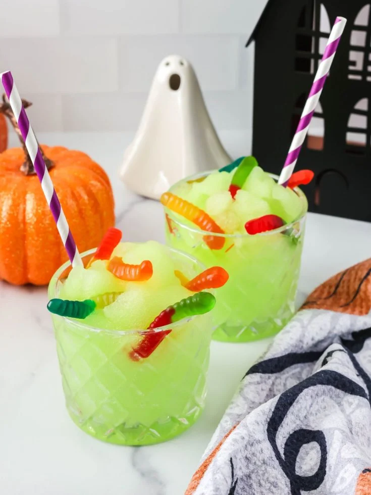 Ninja Slushi witches' brew slush served in two glasses, showcasing its green hue. The slushies are garnished with gummy worms, and Halloween decor is around the glasses.