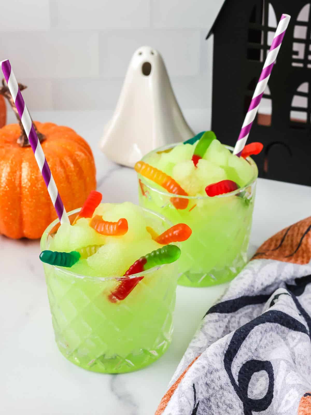 Ninja Slushi witches' brew slush served in two glasses, showcasing its green hue. The slushies are garnished with gummy worms, and Halloween decor is around the glasses.