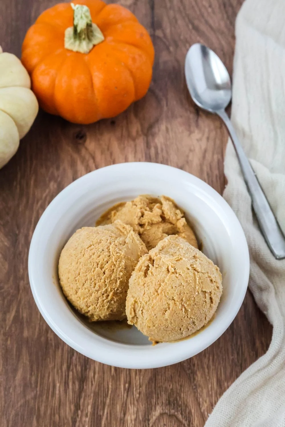 a white bowl serves scoops of Dash Mug pumpkin ice cream. A spoon and mini pumpkins are in the background.