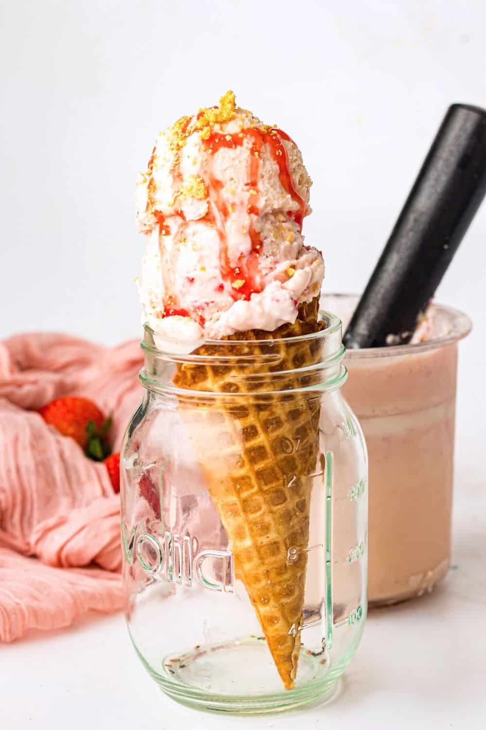 scoops of strawberry cheesecake ice cream served on a waffle cone, supported in a glass jar. The pint of ice cream is in the background.
