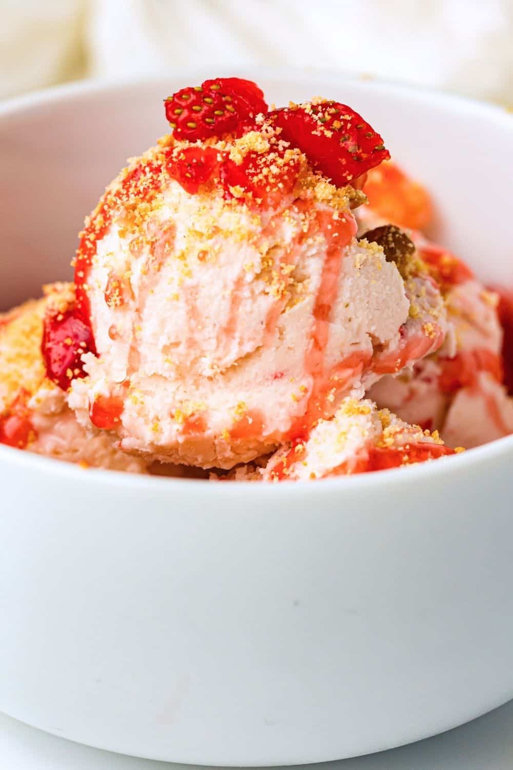 scoops of strawberry cheesecake ice cream in a white bowl, garnished with graham cracker crumbs and diced strawberries.