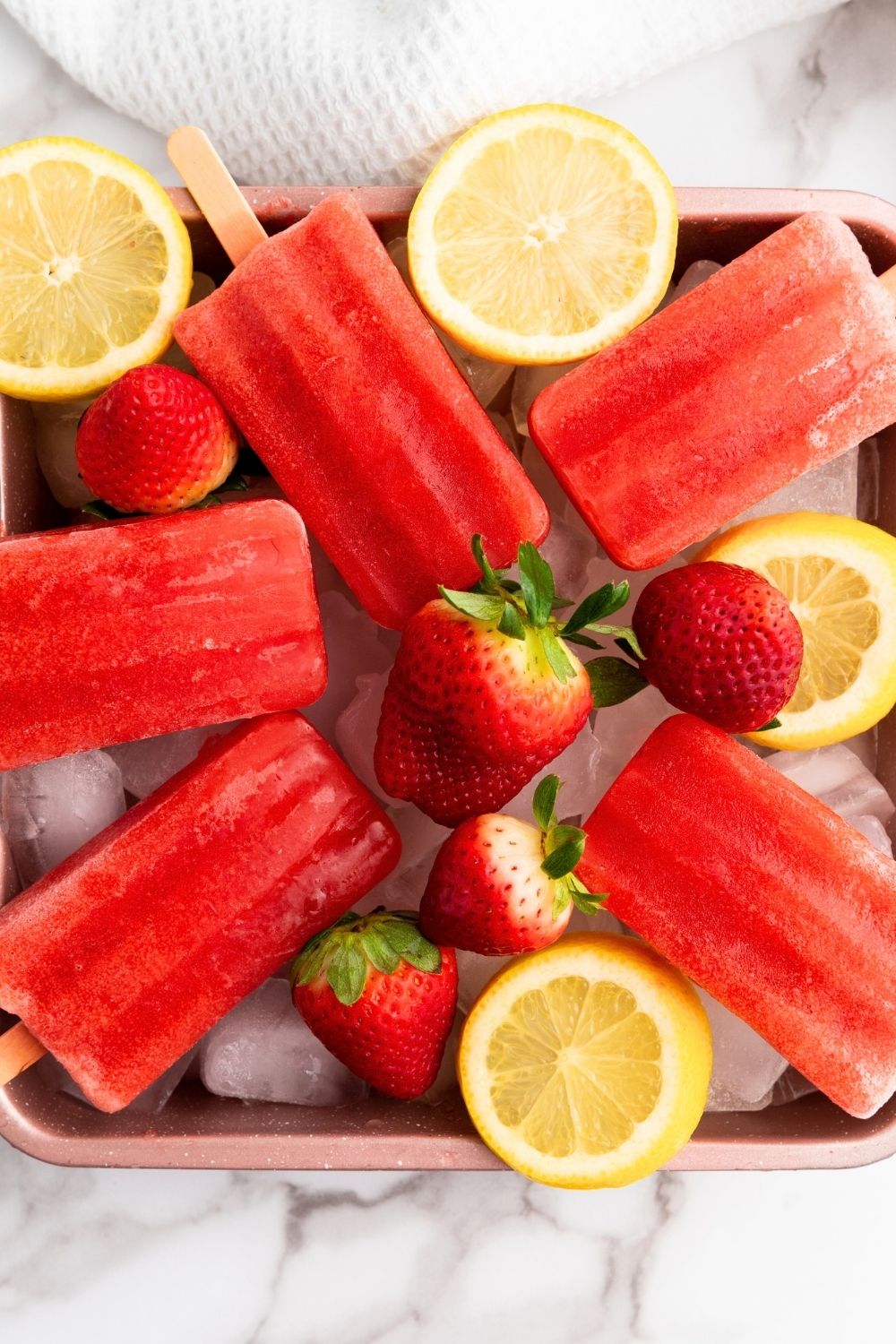 overhead view of a tray serving popsicles made with strawberries and lemon juice. Fresh strawberries and lemon slices garnish the tray.