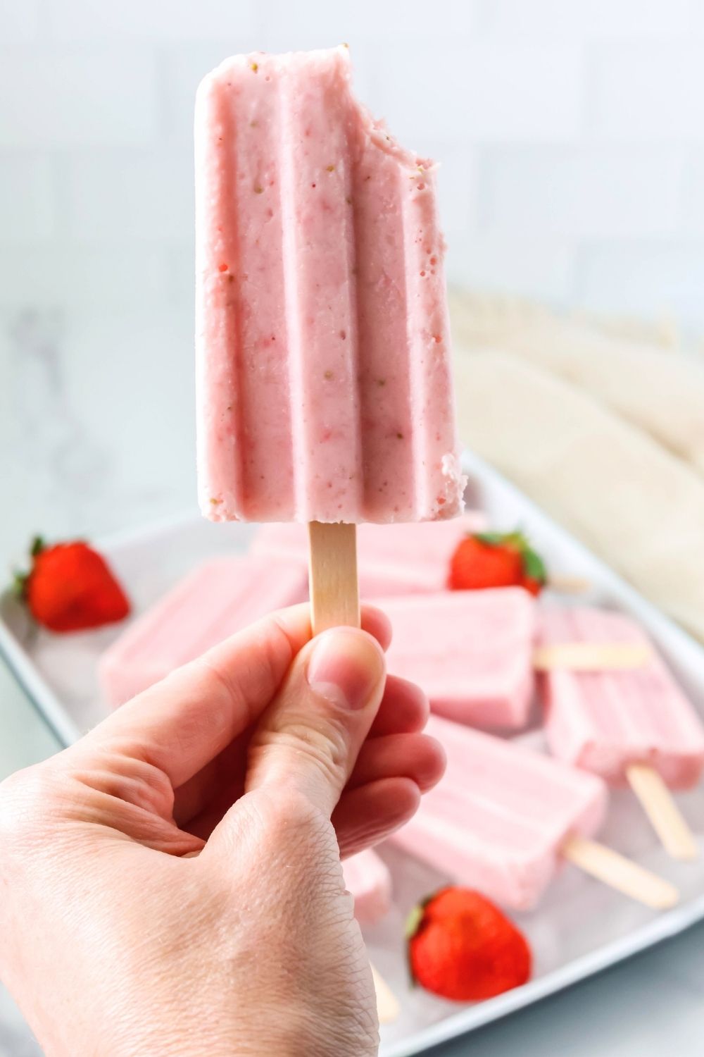 a woman's hand holds a Greek yogurt strawberry popsicle with a bite taken out of it.