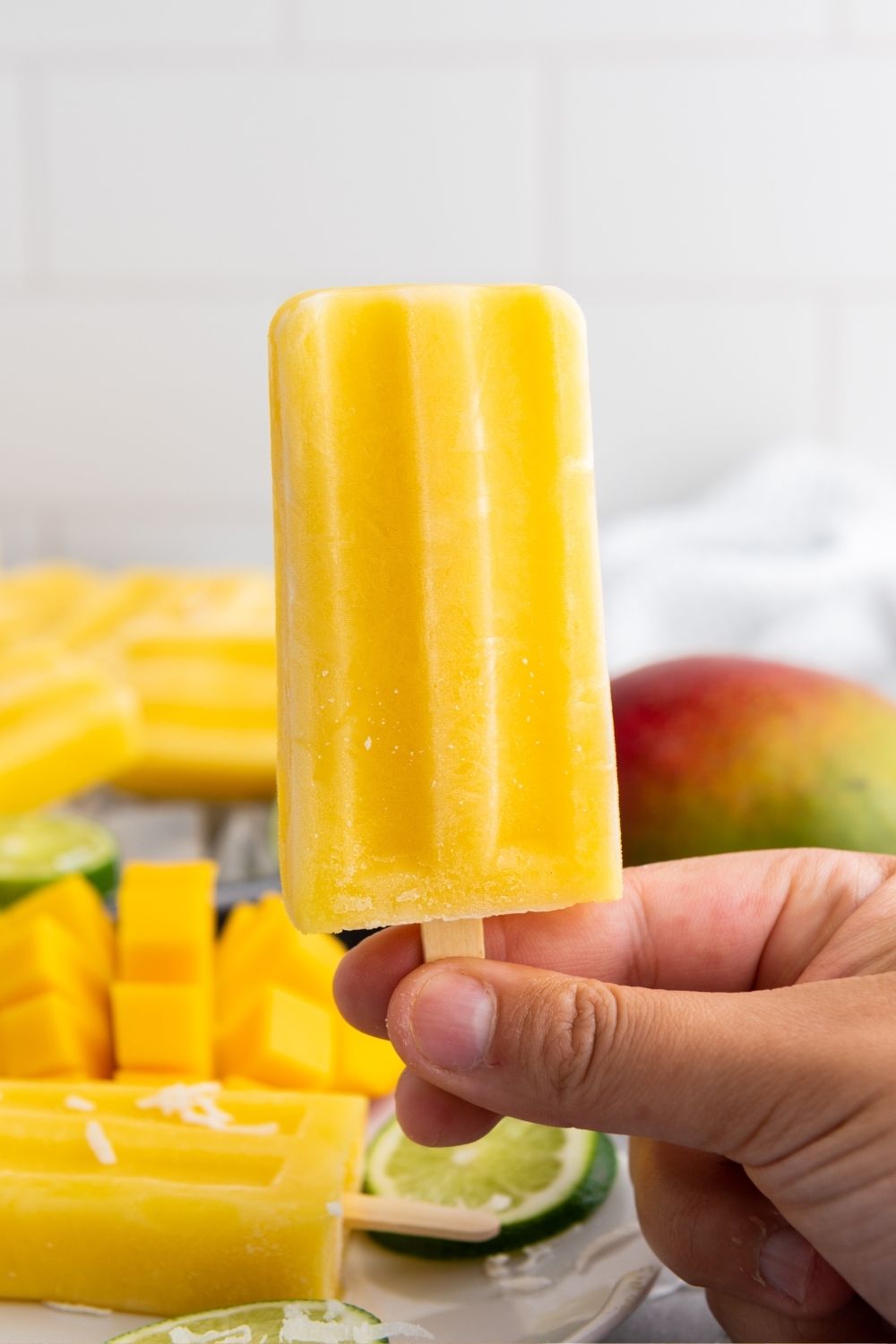 a woman's hand holds a mango popsicle. In the background are more popsicles and mango chunks.