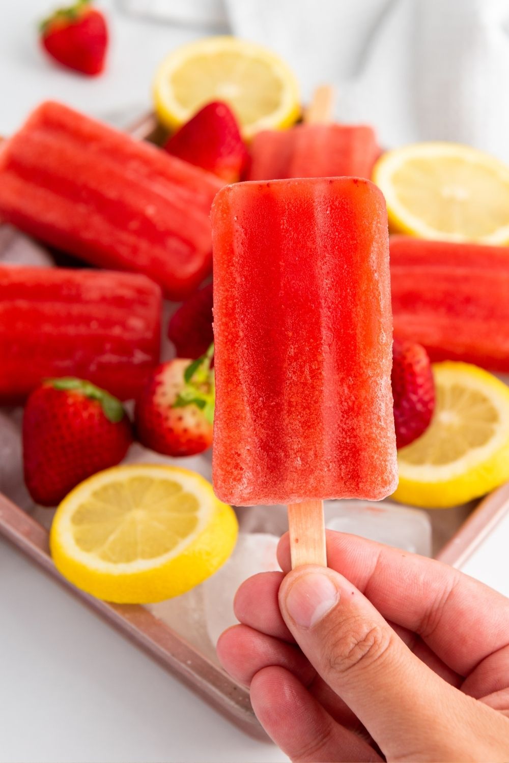 a woman's hand holds a strawberry lemonade paleta, with more popsicles in the background.