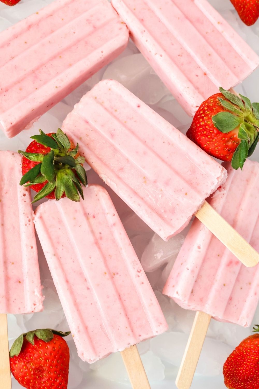 overhead view of higher-protein strawberry yogurt popsicles served on a platter of ice cubes, garnished with fresh strawberries.