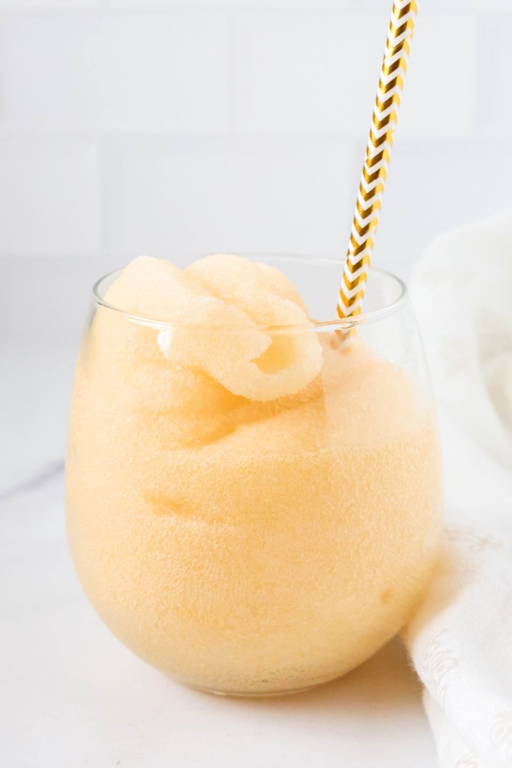 Side view of a glass of peach slushy with a gold and white straw.