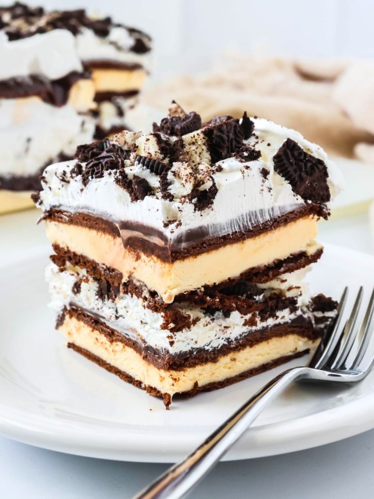 Slice of ice cream sandwich cake, made with hot fudge, Cool Whip, and Oreos, served on a white plate, with a fork next to it.