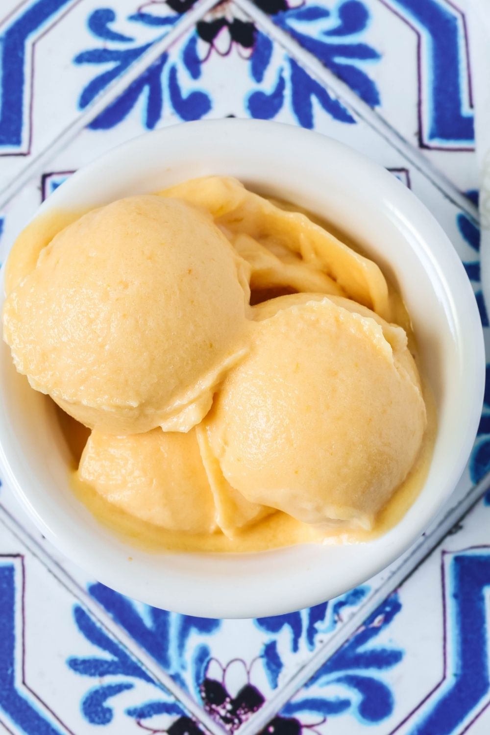 overhead view of scoops of apricot sorbet in a white bowl, on a blue and white tiled background.