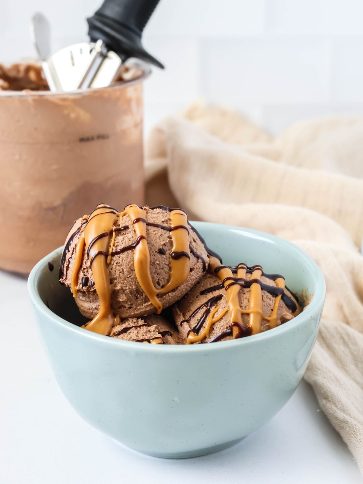 Scoops of Ninja Creami chocolate peanut butter ice cream in a blue bowl, topped with a peanut butter and chocolate drizzle. The Ninja Creami pint container is in the background.