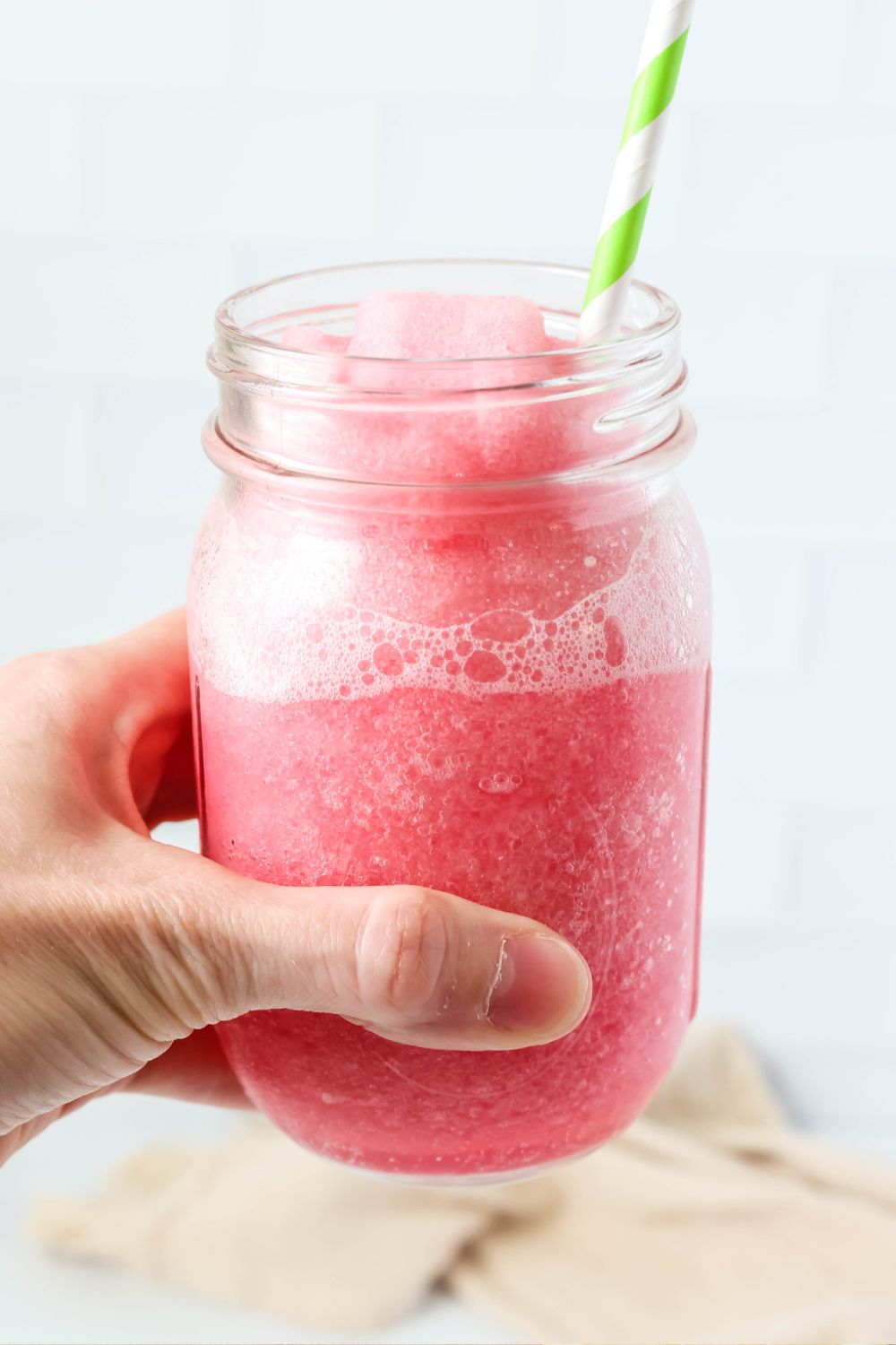 A woman's hand holds a glass jar filled with pink hibiscus limeade made in the Ninja Slushi machine. A straw is in the glass.
