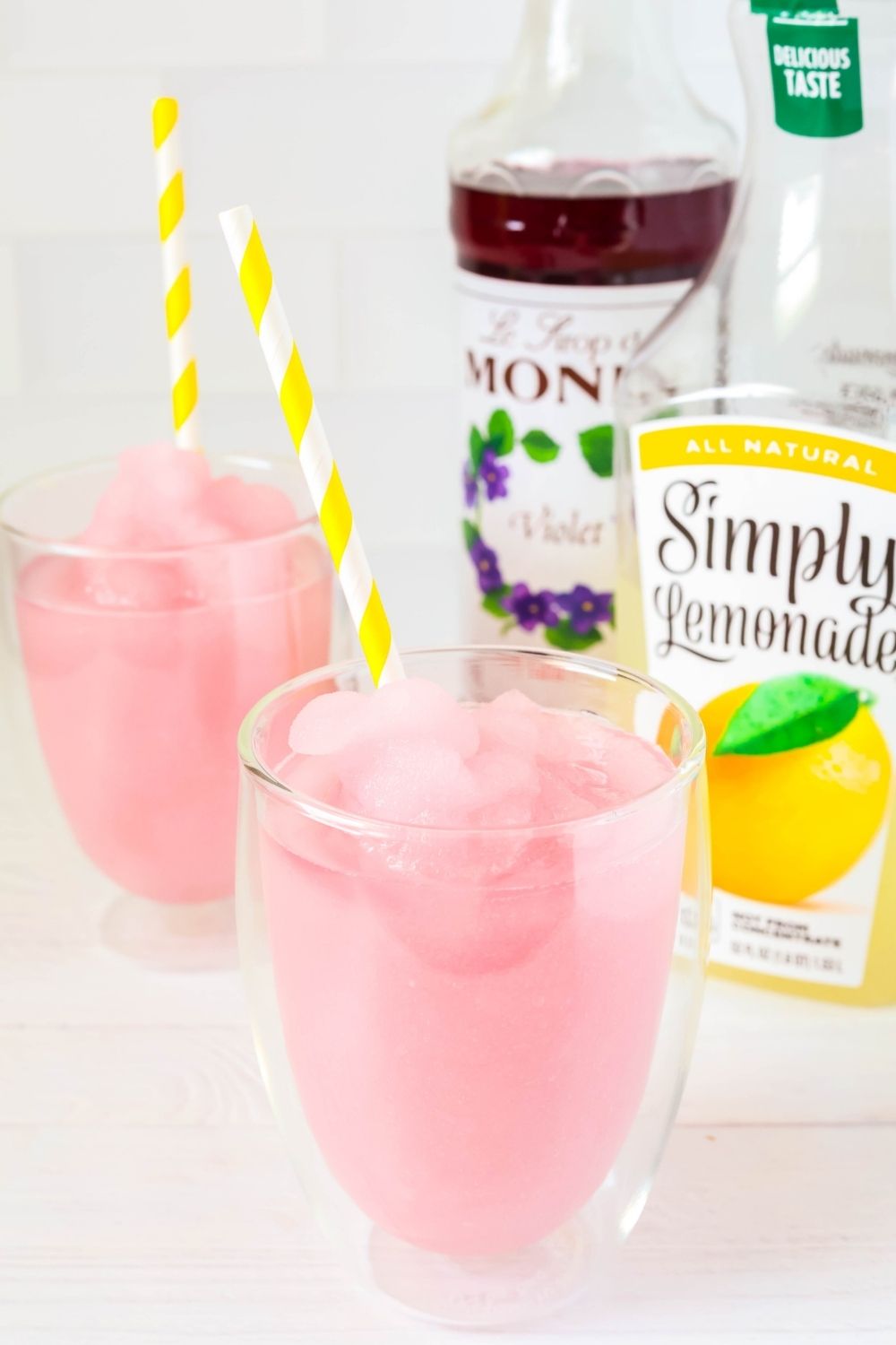Two glasses of violet lemonade slush. A bottle of lemonade and a bottle of violet syrup are in the background.