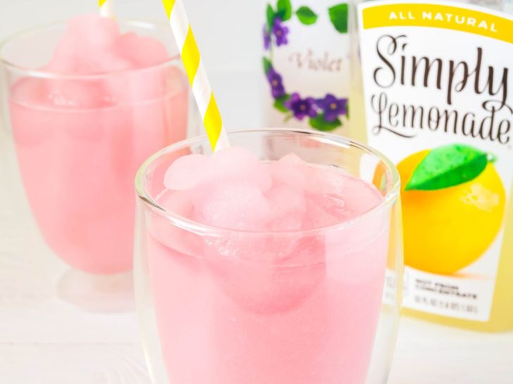 Ninja Slushi violet lemonade slush served in two glasses with yellow and white striped straws. In the background are a bottle of Simply Lemonade and Violet Syrup.