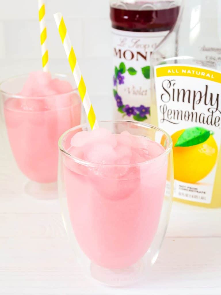 Ninja Slushi violet lemonade slush served in two glasses with yellow and white striped straws. In the background are a bottle of Simply Lemonade and Violet Syrup.